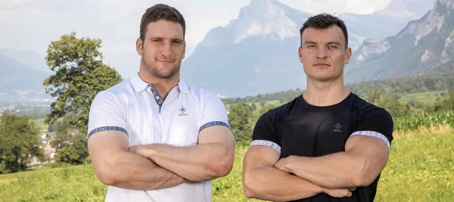 Zwei Männer tragen Edelvetica Shirts in Weiss und Schwarz, posieren mit verschränkten Armen vor imposanter Schweizer Bergkulisse – Symbol für Stärke, Qualität und Swiss Made Mode