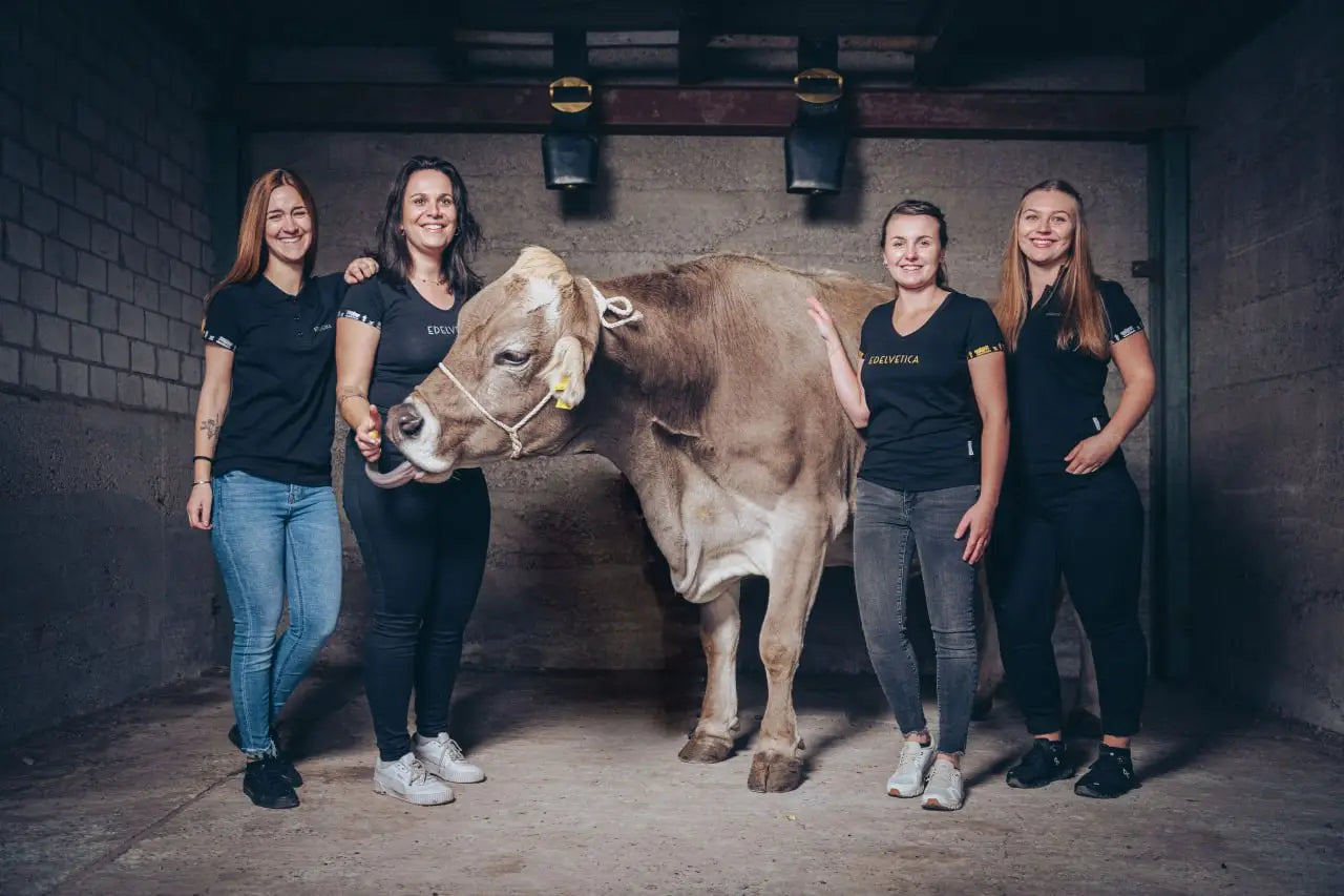 Vier Frauen in schwarzen Edelvetica-Shirts stehen in einem Stall neben einer Braunviehkuh und lächeln in die Kamera.