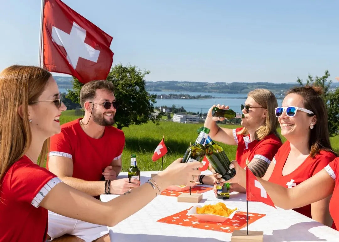 Gruppe von Freunden in roten Shirts stößt am Schweizer Nationalfeiertag mit Bier an, im Hintergrund Schweizer Flagge und Alpenlandschaft