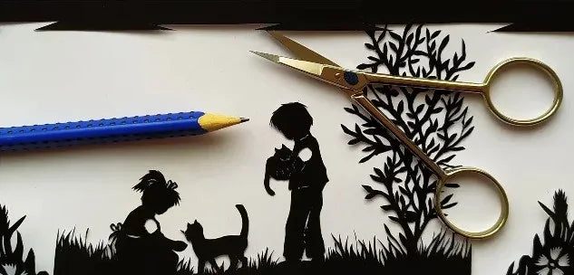 Feiner Scherenschnitt zeigt zwei Kinder mit Katzen und Bäumen, daneben liegen Schere und Bleistift als Werkzeuge der Papercut Kunst
