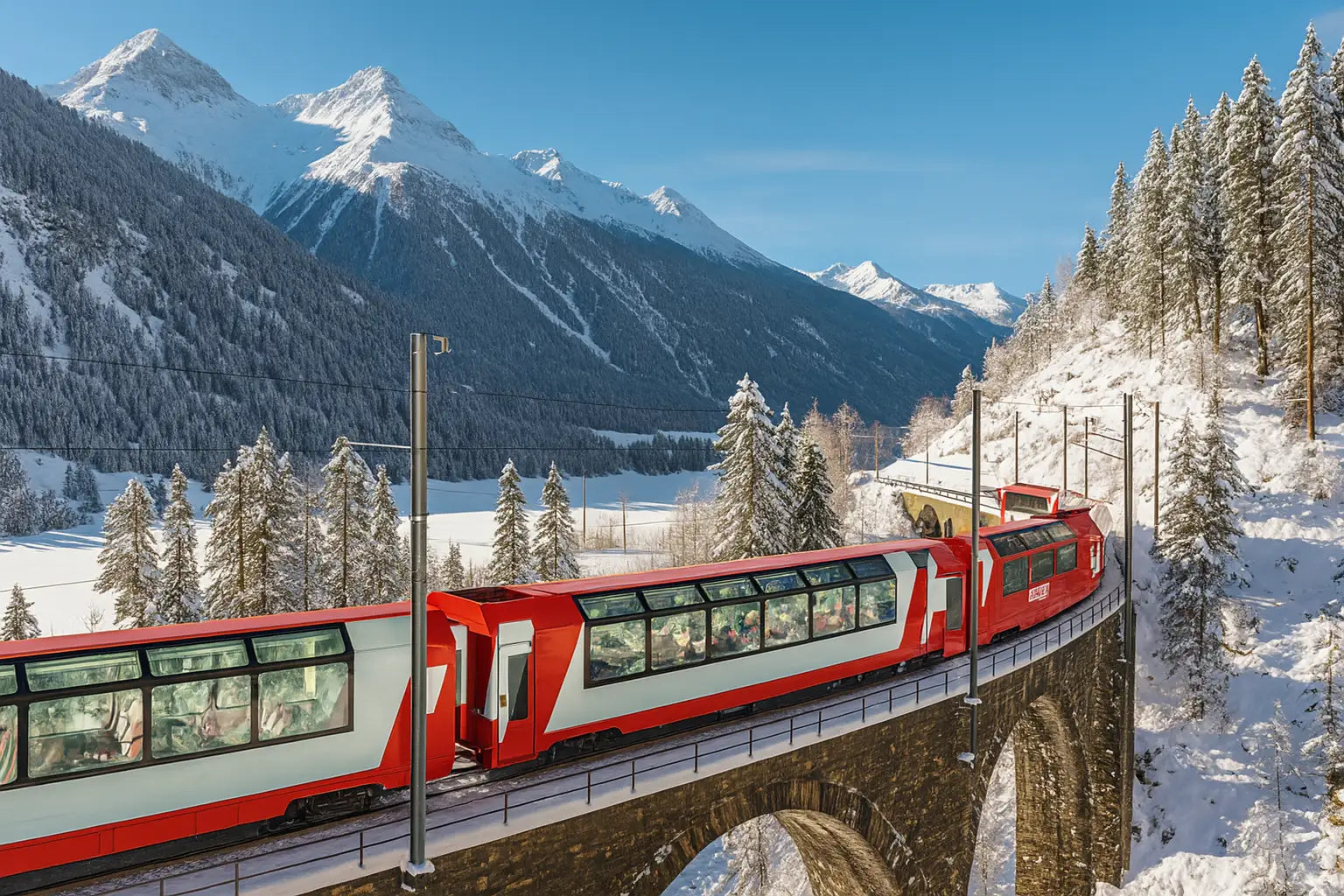 Panoramazug fährt durch tief verschneite Alpenlandschaft mit hohen Gipfeln im Hintergrund.